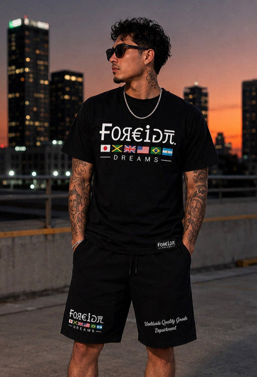 A man wearing a black graphic t-shirt and matching black shorts stands on a rooftop at sunset with a city skyline in the background. The shirt and shorts feature the text 'Foreign Dreams' with multiple colorful flag graphics.