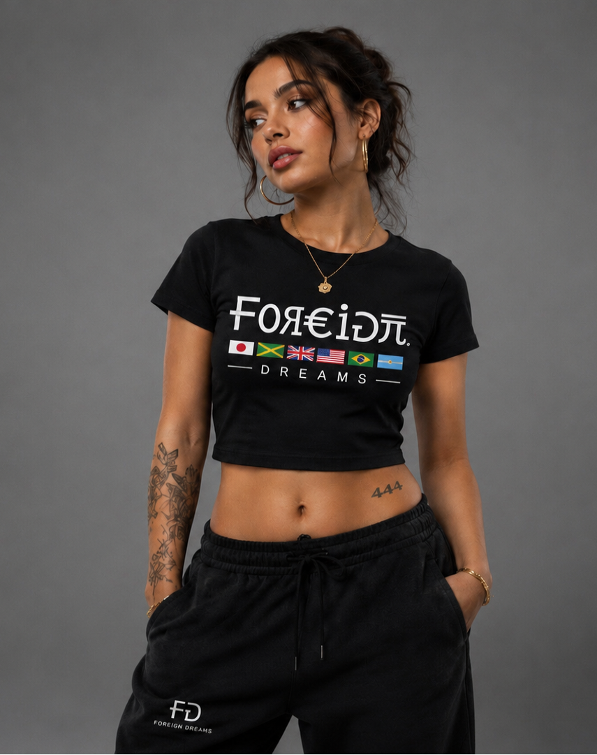A woman modeling a black crop top t-shirt with a Greek flag graphic and text, paired with black sweatpants, against a solid gray studio background.