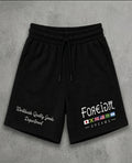 Black sweat shorts with a drawstring waist, featuring white embroidered text on the left leg and a graphic on the right leg with the text 'FOREIDπ. DREAMS' and small colorful flag icons, displayed flat on a gray textured background.