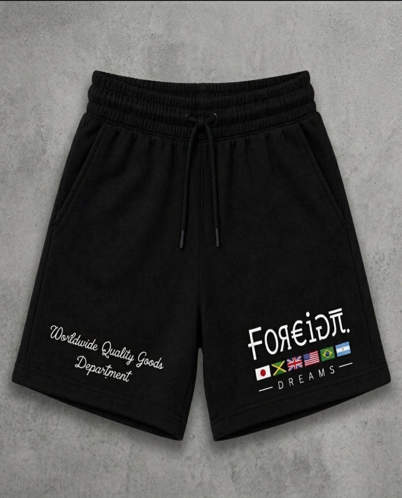 Black sweat shorts with a drawstring waist, featuring white embroidered text on the left leg and a graphic on the right leg with the text 'FOREIDπ. DREAMS' and small colorful flag icons, displayed flat on a gray textured background.