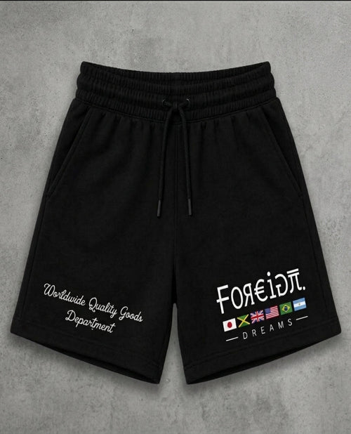 Black sweat shorts with a drawstring waist, featuring white embroidered text on the left leg and a graphic on the right leg with the text 'FOREIDπ. DREAMS' and small colorful flag icons, displayed flat on a gray textured background.