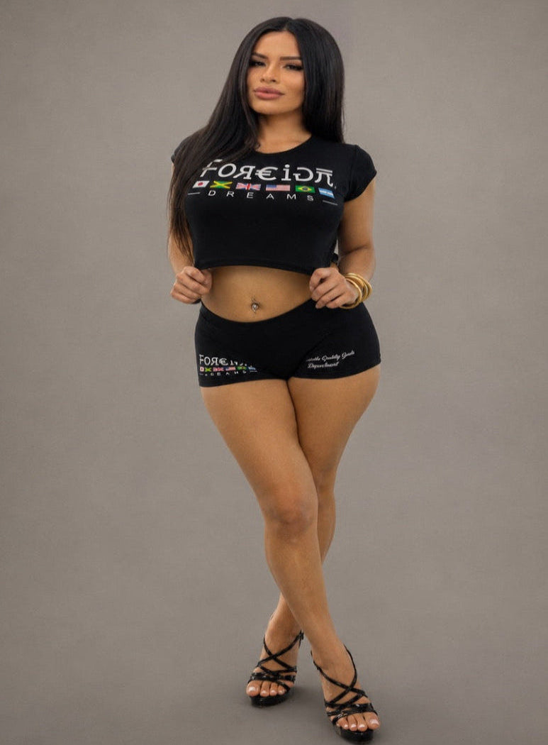 A woman modeling a black short-sleeve crop top and matching black shorts with white and multicolor graphic text, posed against a plain gray studio background.